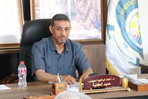 Man in a blue shirt sits at a wooden desk in an office, with a nameplate in Arabic and a flag in the background.