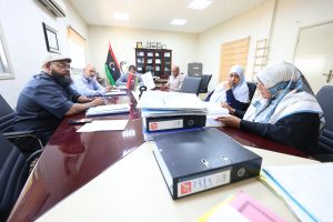 Group of diverse professionals seated around a long conference table with papers and binders in a bright office. Flags are visible in the background.