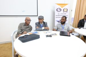 3067EDDD-773A-4994-9658-95177ADC6E00 - جامعة سبها | Sebha University Three men sit around a white round table in a conference room, with documents and a laptop on the table and a banner in the background.
