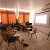 Small seminar in a classroom with five panelists at a desk facing attendees; projection screen displays slides; camera on a tripod nearby.