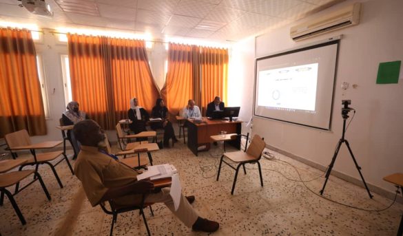 Small seminar in a classroom with five panelists at a desk facing attendees; projection screen displays slides; camera on a tripod nearby.