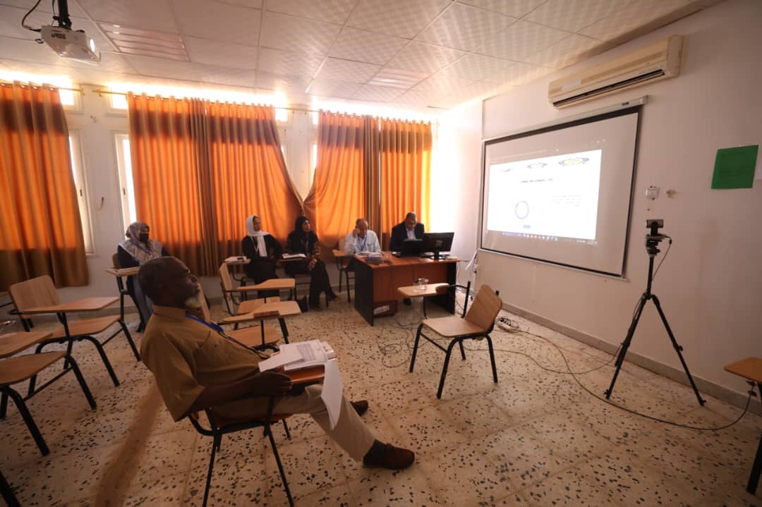 Small seminar in a classroom with five panelists at a desk facing attendees; projection screen displays slides; camera on a tripod nearby.