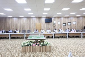 Conference room meeting with professionals seated along a long table; floral centerpiece in foreground and wall-mounted certificates behind.