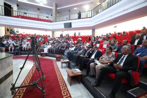 34F9AD84-2C65-43D3-9AF1-3018AB7AF926 - جامعة سبها | Sebha University Formal audience seated in red theater-style chairs for a conference or ceremony; camera on a tripod in the foreground facing the stage.