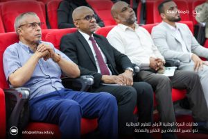 3EE56C73-A992-4912-9C11-9685A9B6A5B8 - جامعة سبها | Sebha University Four men in business attire sit in red theater seats at a conference, with Arabic caption and a university logo in the corner.