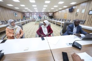 41F245F6-C997-4FEA-8FE4-100D50E1CD0C - جامعة سبها | Sebha University Three professionals sit at a large conference table in a wood-paneled meeting room with flags displayed in the background.