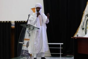 43365D02-E4B0-4CFE-9DE9-E10B2438C598 - جامعة سبها | Sebha University Man in a white traditional robe and turban speaks into a microphone at a glass lectern on a stage with dark curtains behind him.