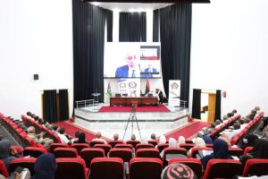 438DCC4B-77E7-471F-A595-206E20D7D508 - جامعة سبها | Sebha University Panel discussion on stage in an auditorium with a large video screen showing a speaker and banners beside the stage.
