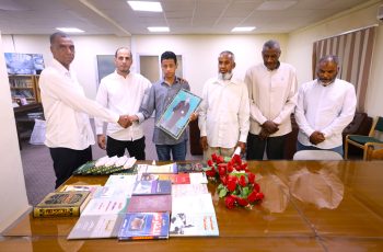 Seven people, six men in white shirts and a teenage boy in a striped shirt, stand around a table of books in a meeting room; the boy holds a framed photograph.