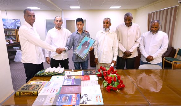 4541A007-A69B-4D5D-98A7-0C661004FD68 - جامعة سبها | Sebha University Seven people, six men in white shirts and a teenage boy in a striped shirt, stand around a table of books in a meeting room; the boy holds a framed photograph.