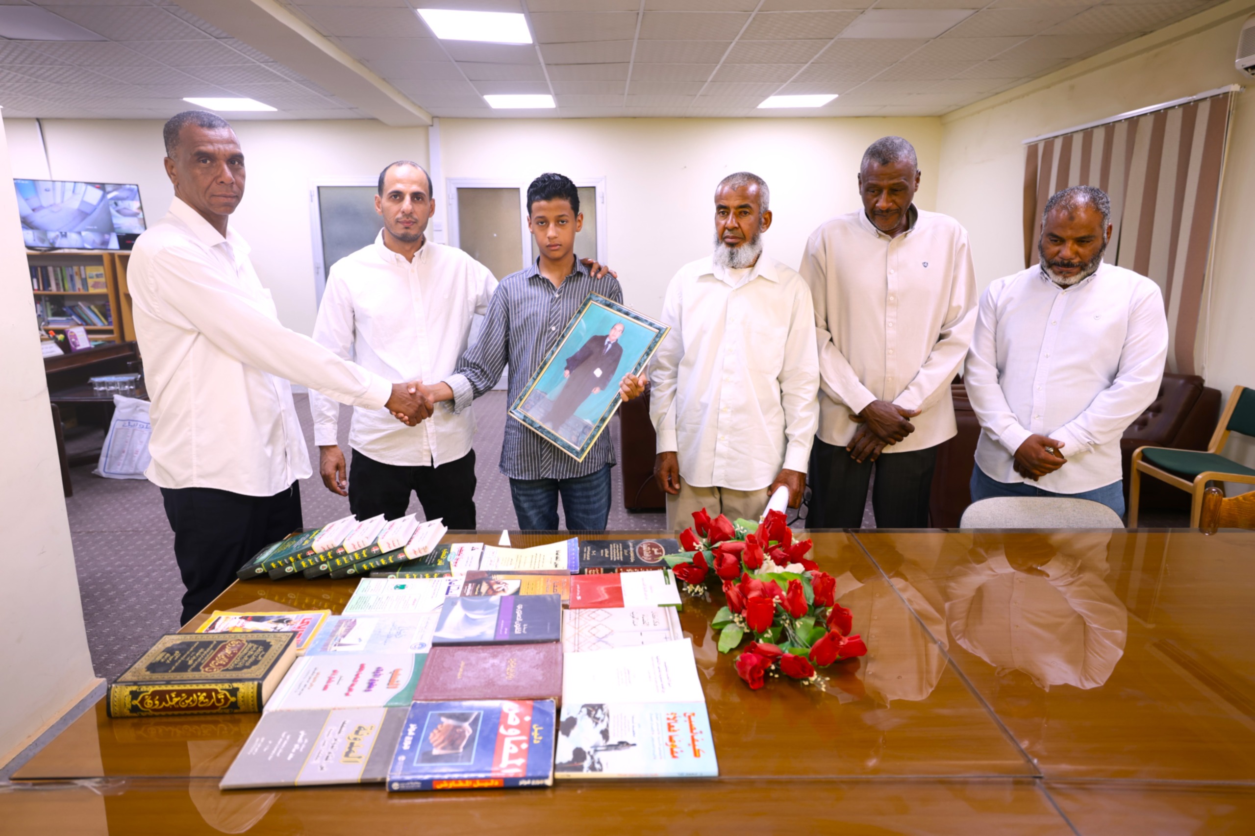 Seven people, six men in white shirts and a teenage boy in a striped shirt, stand around a table of books in a meeting room; the boy holds a framed photograph.