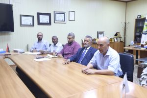457E8DEC-017D-4361-82B2-82DC31830AD9 - جامعة سبها | Sebha University Group of men sit around a wooden conference table in a formal meeting room, taking notes and talking