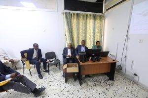 4689B4C5-FB4E-4252-9045-37F682C98C4B - جامعة سبها | Sebha University Five men in business attire sit along and around a wooden desk in a small meeting room, with laptops and a projector setup for a presentation.