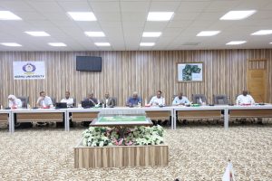 Conference room at Sebha University with people seated around a long U-shaped table and a banner reading 'SEBHA UNIVERSITY' on the wall behind them.