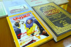 Colorful magazines on a wooden table; front cover shows a woman in yellow pouring tea, with a yellow border, beside a large ornate black-and-gold book.