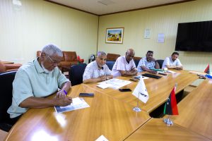 49CBF2B4-E3E3-4D7B-B490-39EC61789D22 - جامعة سبها | Sebha University Group of men seated around a long conference table reviewing documents in a meeting room, small flags on the table and a large screen on the wall.
