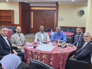 Six professionals in suits sit around a round table with papers and water bottles in a formal meeting room.