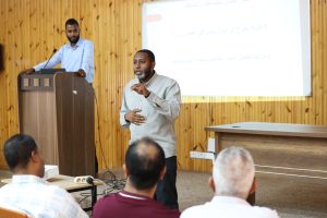 Speaker at a wooden lectern with a microphone, presenting to an audience in a room with a projector screen showing Arabic text behind him.
