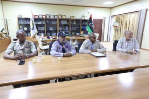 Four men sit at a long wooden conference table in a meeting room, one in uniform, another in scout-like attire, with Libyan flags and shelves of plaques behind them (informative scene).