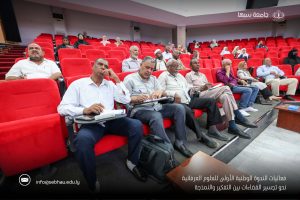 Audience in a red-upholstered lecture hall listening and taking notes, with Arabic text and logos overlayed on the image's bottom edge.