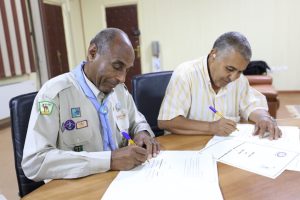 Two men sit at a wooden table signing documents; one wears a badge-filled uniform while the other wears a striped shirt.