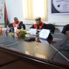 Two men in red academic gowns sit at a conference table with nameplates and a laptop in a meeting room.