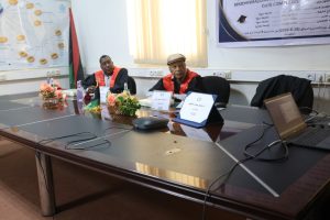 Two men in red academic gowns sit at a conference table with nameplates and a laptop in a meeting room.