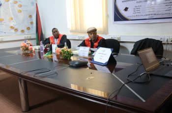 Two men in red academic gowns sit at a conference table with nameplates and a laptop in a meeting room.