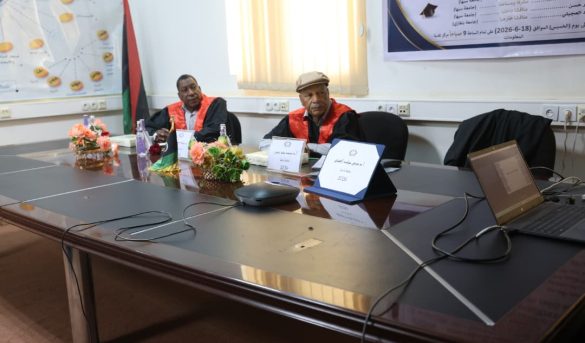 58B382D1-CB52-4E44-8866-8A82041F5B15 - جامعة سبها | Sebha University Two men in red academic gowns sit at a conference table with nameplates and a laptop in a meeting room.