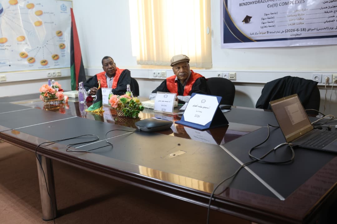 Two men in red academic gowns sit at a conference table with nameplates and a laptop in a meeting room.