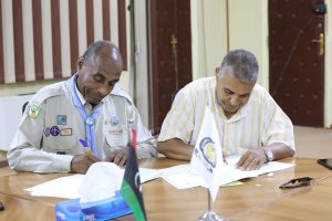 Two men sign documents at a wooden table during a formal signing ceremony, with flags and tissue box nearby.