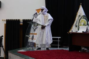 5AC85489-D8E4-48A7-9694-39F722BCA2FF - جامعة سبها | Sebha University Man in traditional white attire and turban stands at a clear podium on a stage, addressing an audience with banners and a flag nearby.