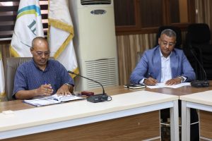 Two men at a conference table signing documents; microphones, notebooks, and flags behind them.