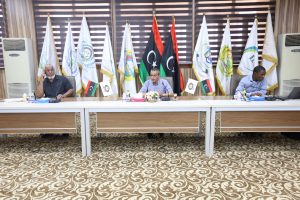 Three men sit at a long conference table with microphones and water bottles, flags lined up behind them.