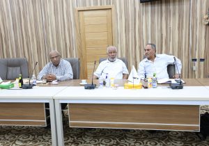 Three men sit at a long conference table in a wood-paneled room, with bottles and papers in front, appearing to discuss anew meeting.