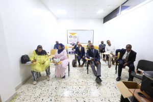 5E19005B-DE15-4AB7-98AF-C5809D92DF8A - جامعة سبها | Sebha University Group of professionals seated in a bright, white-walled training room, waiting for a session with notebooks and bags nearby.
