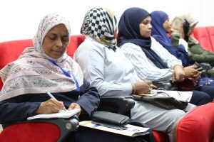 Female attendee in a floral hijab writing in a notebook, with others seated in red chairs behind her.