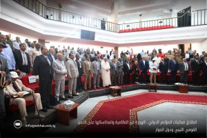 Group of formally dressed people standing in a circular, tiered conference hall with red seats during an opening ceremony. A banner caption in Arabic runs along the bottom edge of the image.