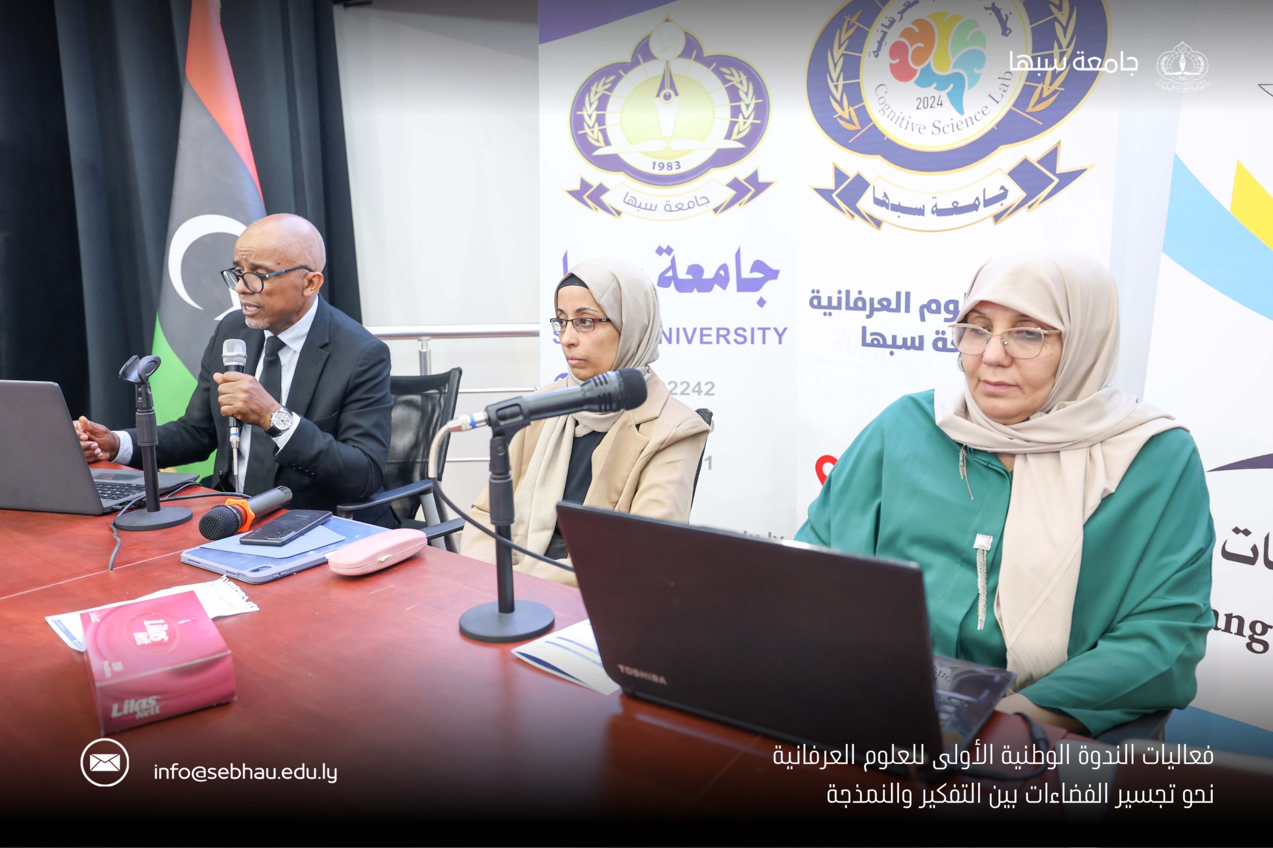 Panelists seated at a table with microphones and laptops during a university event, banners with logos in the background.