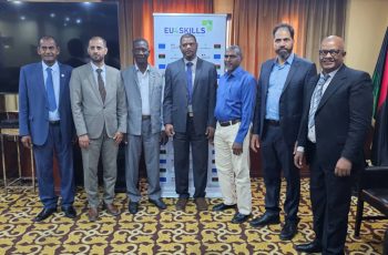 Seven men in suits standing in a row at an EU4Skills event, posing for a group photo indoors in front of a banner.