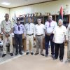 Eight men stand in an office, several in scout-like uniforms with badges and neckerchiefs, with a flag and trophy shelf in view.