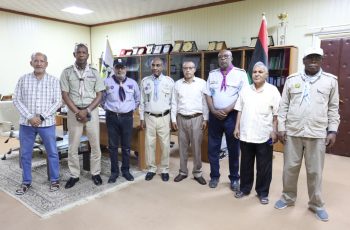 Eight men stand in an office, several in scout-like uniforms with badges and neckerchiefs, with a flag and trophy shelf in view.