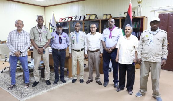 Eight men stand in an office, several in scout-like uniforms with badges and neckerchiefs, with a flag and trophy shelf in view.