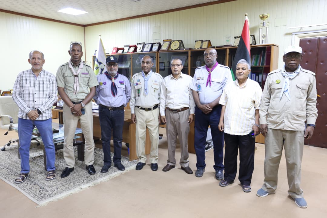 Eight men stand in an office, several in scout-like uniforms with badges and neckerchiefs, with a flag and trophy shelf in view.