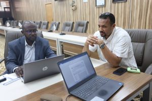 Two men sit at a conference table with open laptops, discussing a project.