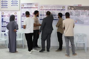 Group of people standing and reading large posters with Arabic text pinned to a wall, beside arranged white chairs in a hall.