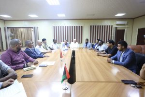 638B5E5E-BF55-49E7-A4E7-E7AB8F87F061 - جامعة سبها | Sebha University People sit around a large conference table in a meeting room; small flags on the table.