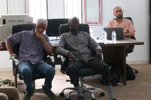 64C8CE3E-9B00-4C6F-B6D8-242525117856 - جامعة سبها | Sebha University Three men in an office: two sit in front in swivel chairs, while a third sits at a desk with a laptop and water bottles behind them.
