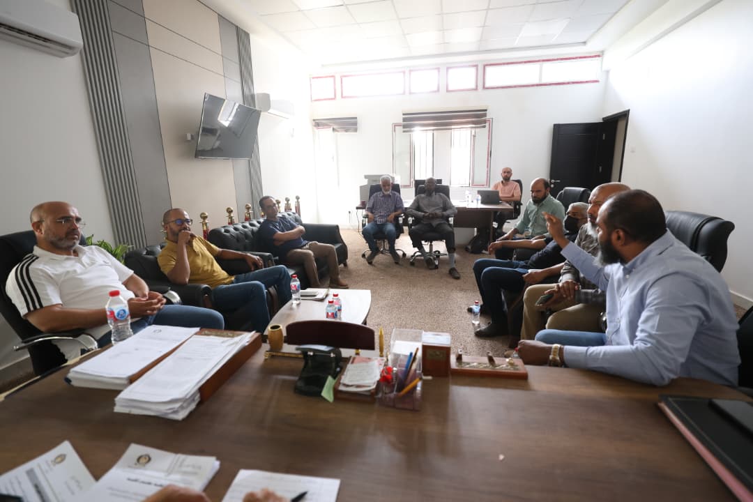 Group of men in a meeting room sitting in a semi-circle around a large desk with documents and water bottles.