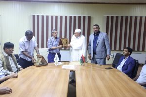 678E9039-4F55-4D69-9D9B-7C6A2850930E - جامعة سبها | Sebha University Men in a conference room exchange plaques during an award ceremony around a long wooden table with flags on it